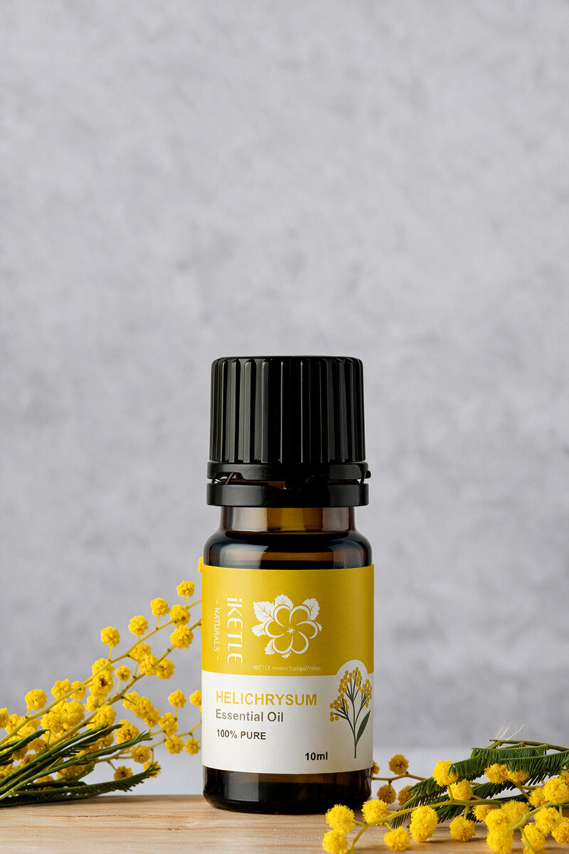 A bottle of iKETLE Helichrysum essential oil beside fresh yellow helichrysum flowers